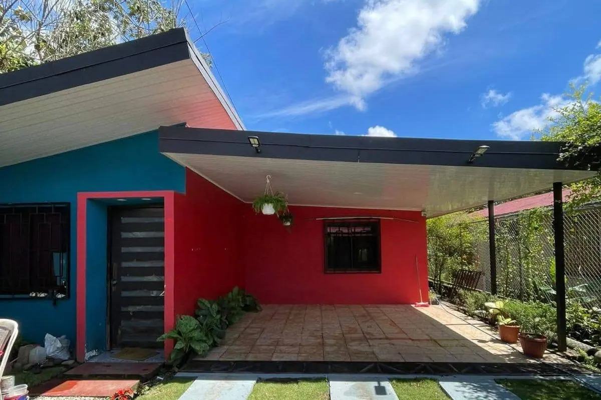 Casa de paredes azul y roja con corredor techado y caminito de concreto entre el zacate